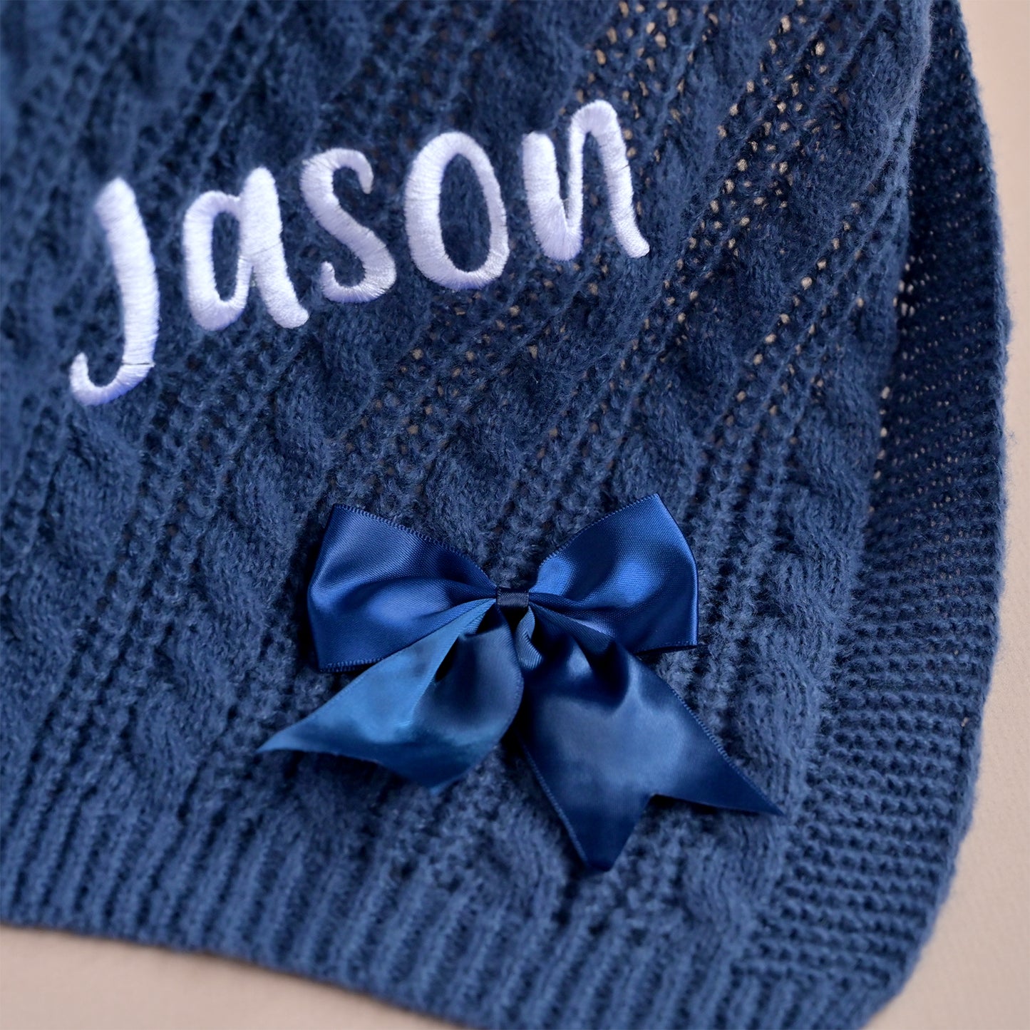 Close-up of a navy blue cable-knit garment, likely a hat or scarf, with the name 'Jason' embroidered in white script and a shiny navy blue bow attached near the bottom.