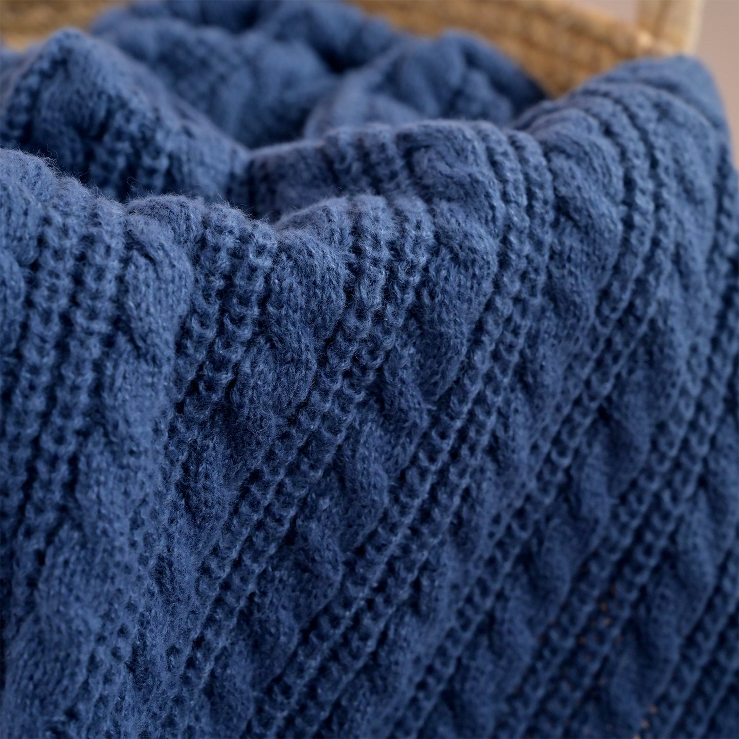 A close-up of a chunky navy blue cable-knit blanket draped over a light brown woven basket.