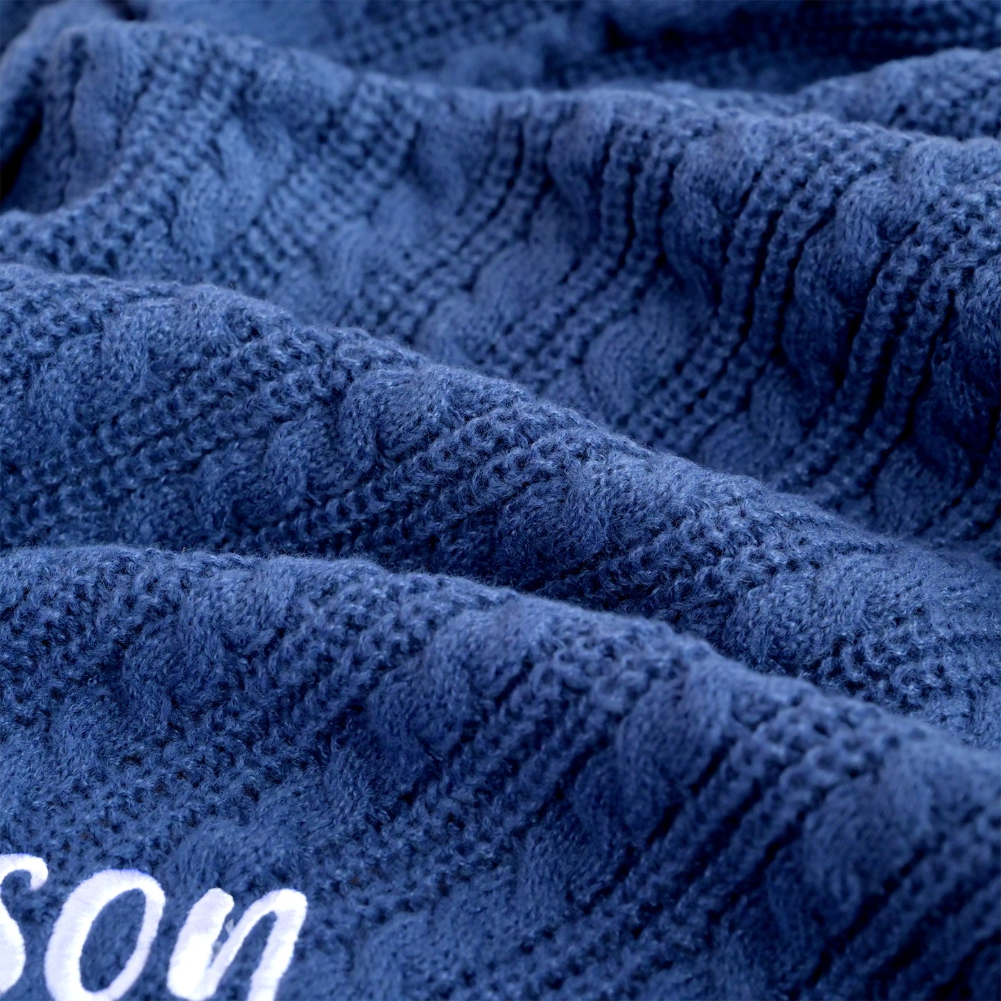 Close-up of a textured navy blue knit fabric with a cable pattern and white embroidered lettering partially visible in the lower left corner.