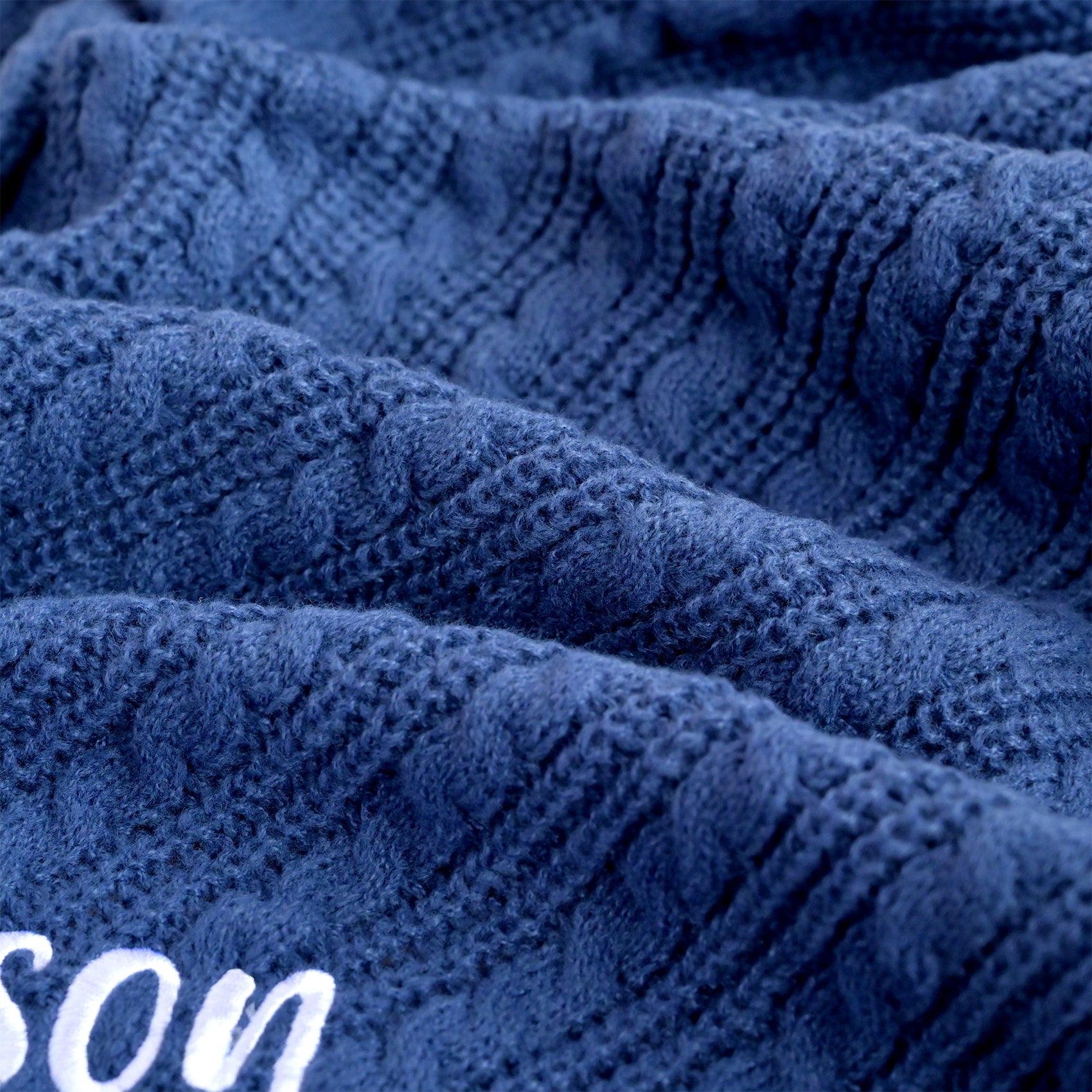 Close-up of a textured navy blue knit fabric with a cable pattern and white embroidered lettering partially visible in the lower left corner.