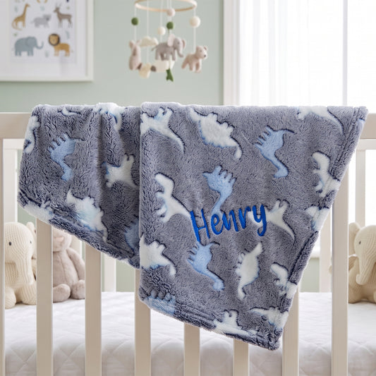 A personalized baby blanket draped over a white crib, featuring a gray plush background with light blue and white dinosaur patterns and the name 'Henry' embroidered in blue.