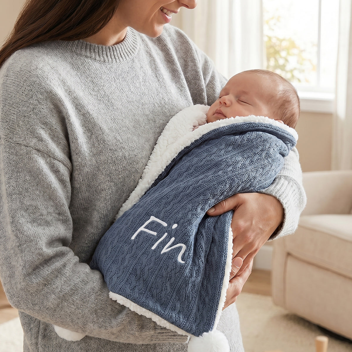 Dark blue cable-knit baby blanket with sherpa fleece lining and pom-pom trim, personalised with the name Fin embroidered in white script, swaddling a sleeping newborn.