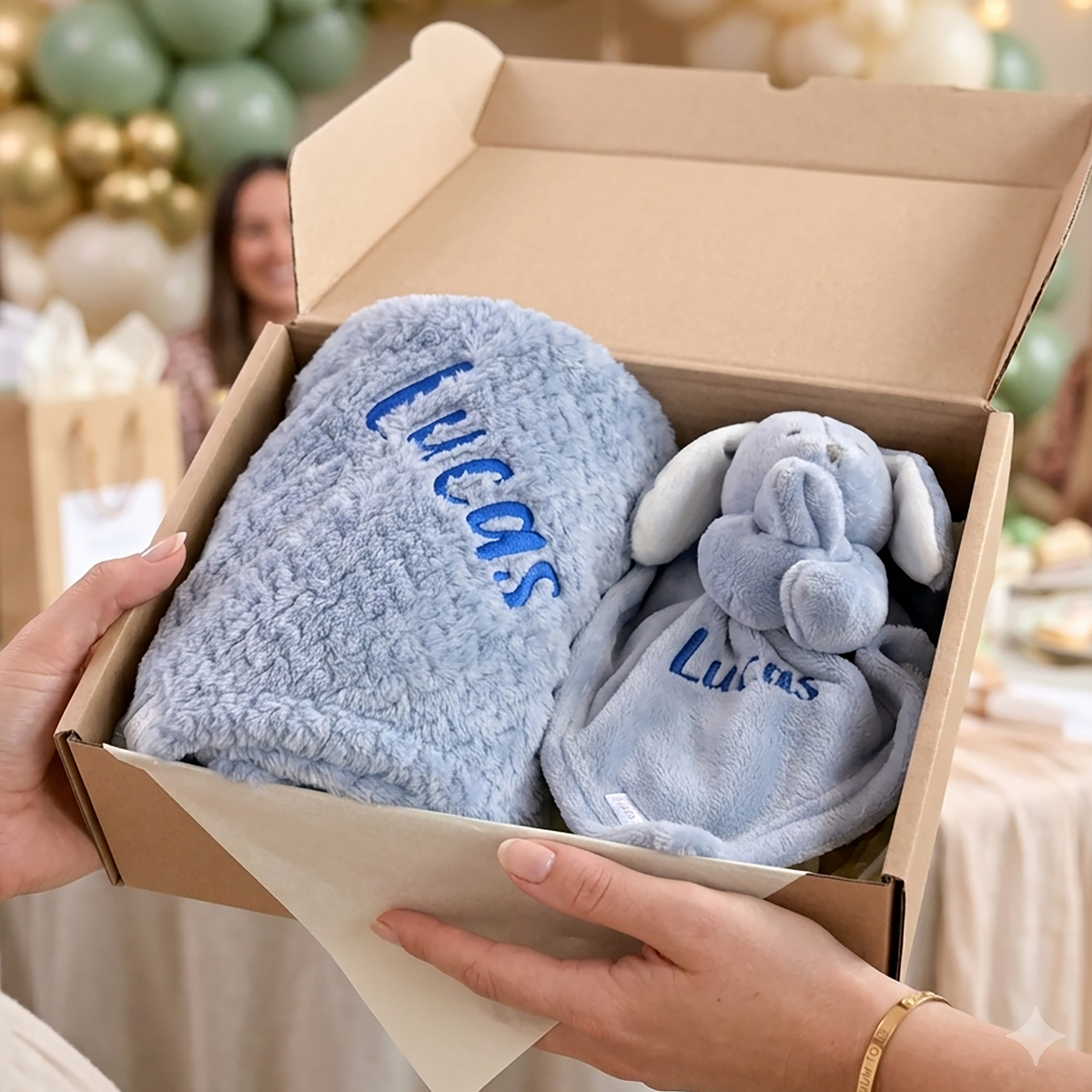 A personalized baby gift set in a kraft paper box, featuring a plush light blue blanket and matching elephant-shaped baby toy, both embroidered with the name 'Lucas' in blue. Hands are holding the open box, with a blurred party background showing balloons and a woman.