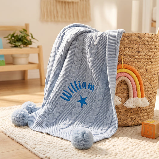 Pale blue cable knit baby blanket with pom-pom corners, personalised with the name William arched above an embroidered star, resting against a woven basket.