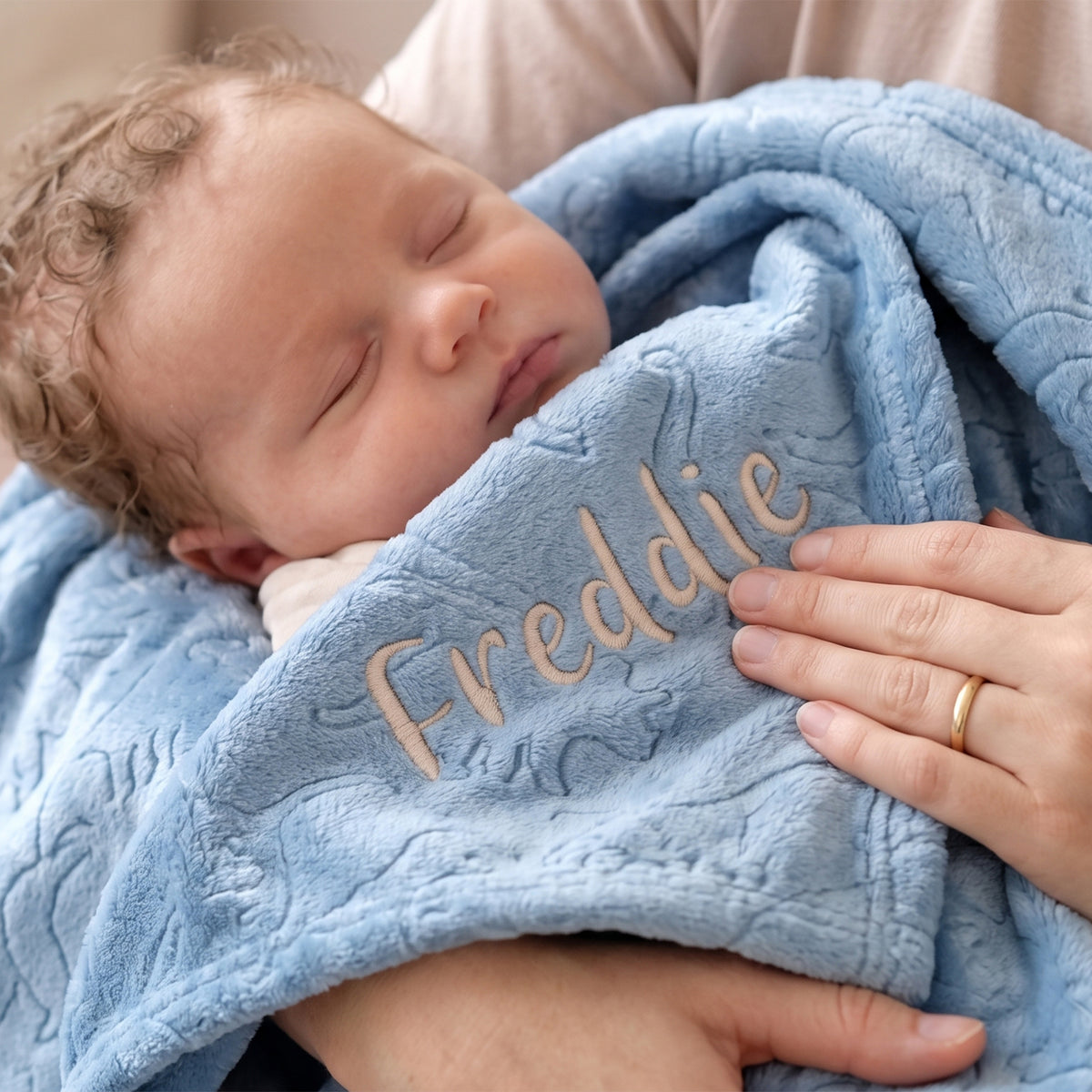 A close-up of a sleeping baby wrapped in a light blue textured blanket with the name 'Freddie' embroidered in beige on the front, held by an adult's hands.