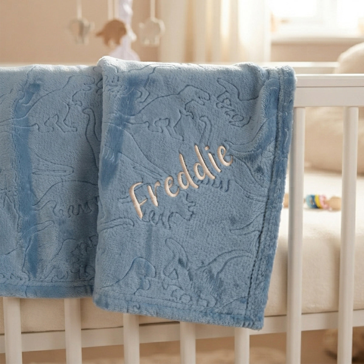 A personalized baby blanket in dusty blue with embossed dinosaur patterns and the name 'Freddie' embroidered in gold, draped over a white crib.