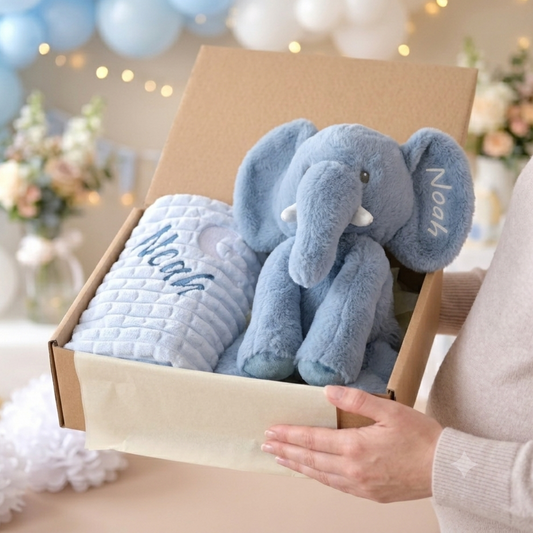 A person holds an open cardboard gift box containing a light blue plush elephant toy and a matching light blue quilted blanket, both personalized with the name 'Noah' in white script. The background shows a softly blurred party setting with balloons and string lights.