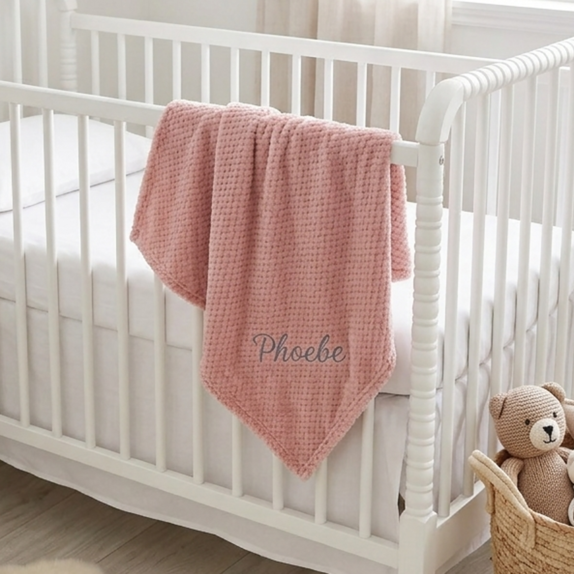 Dusty pink honeycomb fleece baby blanket personalised with the name Phoebe in grey embroidery, draped over a white crib beside a knitted teddy bear.