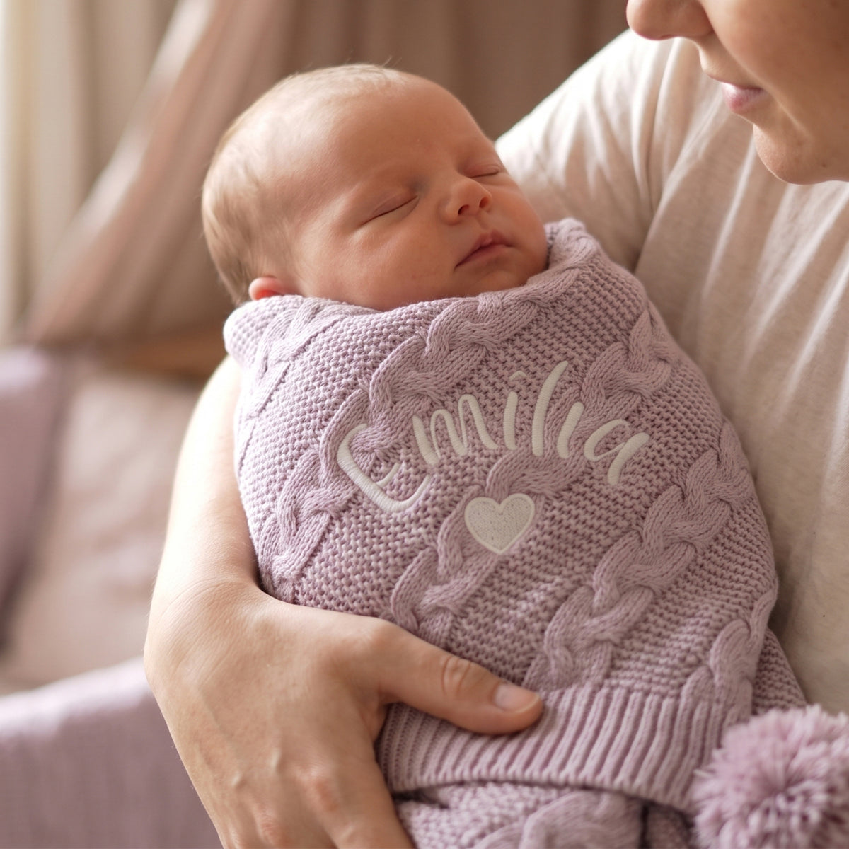 A sleeping newborn baby swaddled in a light pink cable-knit blanket with the name 'Familia' and a heart embroidered on the front, held by an adult in a soft indoor setting.