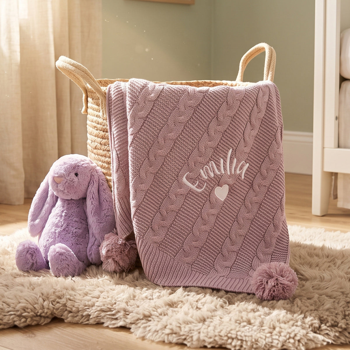 A dusty pink cable-knit baby blanket with the name 'Emma' and a small heart embroidered on the front, draped over a woven storage basket. A purple plush bunny toy sits beside it on a cream shag rug in a softly lit nursery setting.
