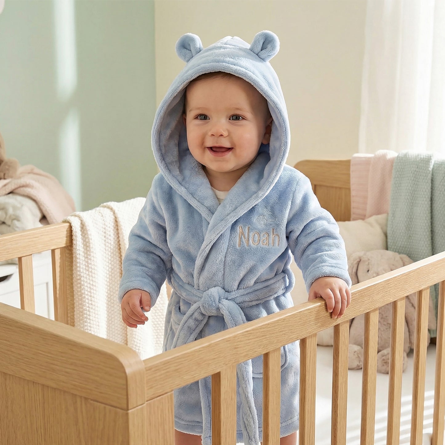 A baby wearing a light blue plush hooded bathrobe with bear ears and the name 'Noah' embroidered on the chest, standing in a wooden crib in a softly lit nursery.