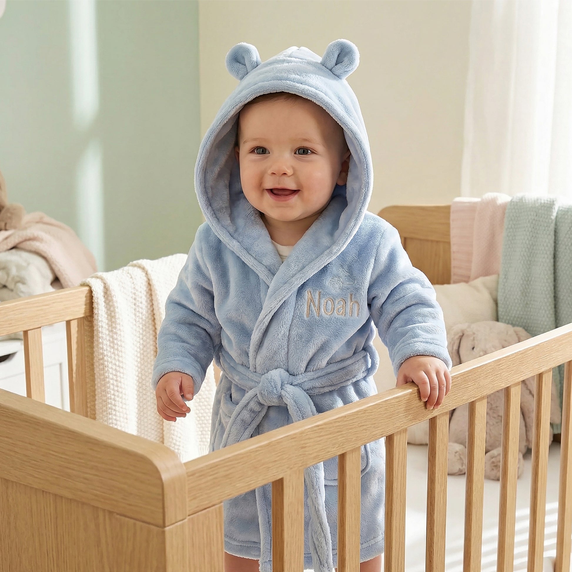 A baby wearing a light blue plush hooded bathrobe with bear ears and the name 'Noah' embroidered on the chest, standing in a wooden crib in a softly lit nursery.