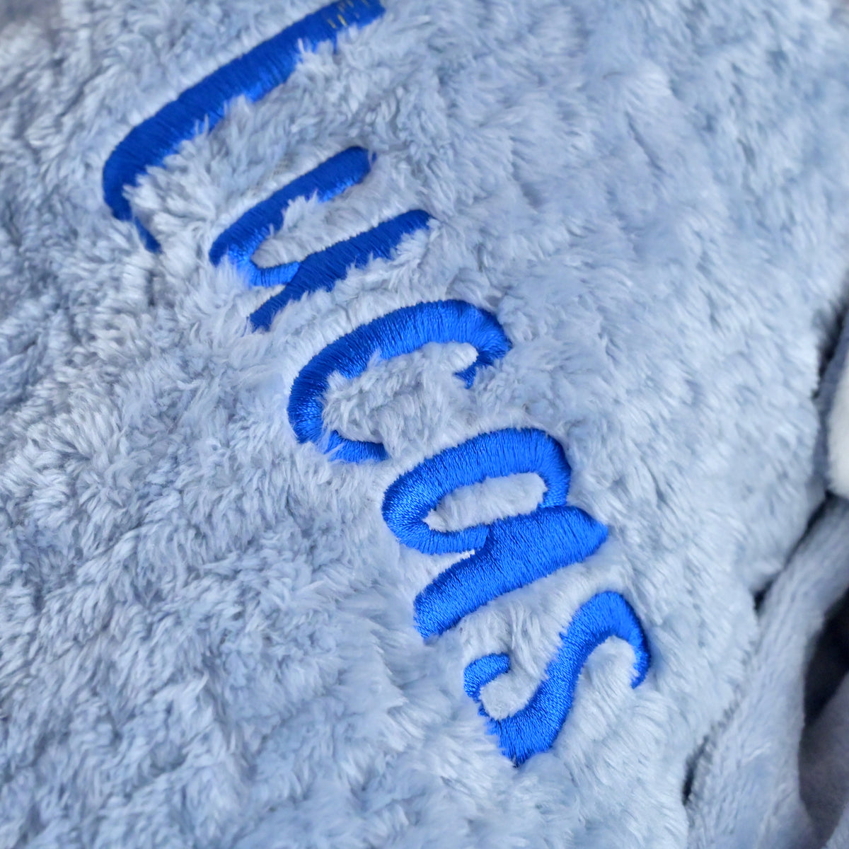 Close-up of a plush light blue-gray fabric with bright blue embroidered lettering running diagonally across the surface.