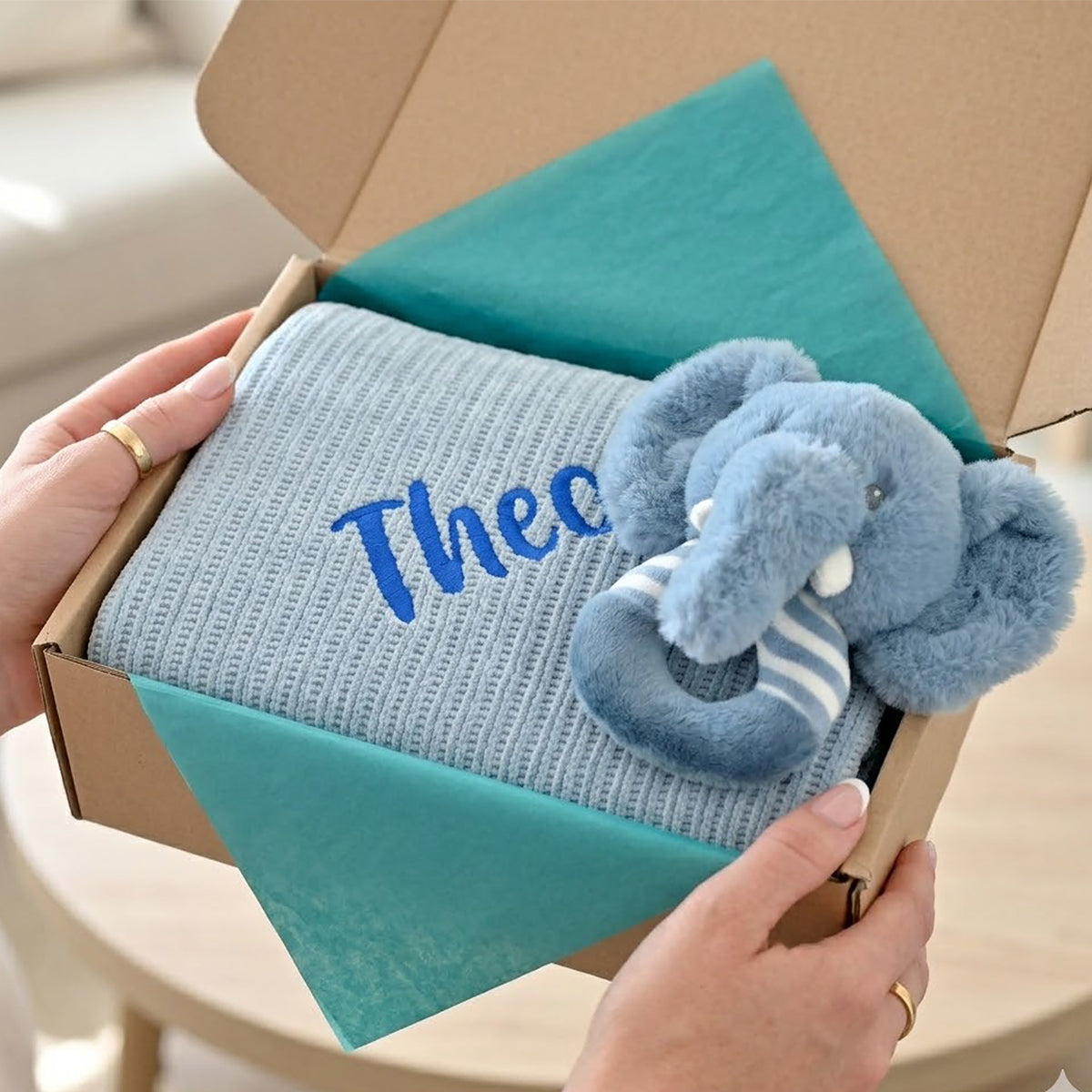 A light blue ribbed blanket with the name 'Thee' embroidered in blue, presented in a cardboard gift box with teal tissue paper and a plush blue elephant toy, held by two hands.