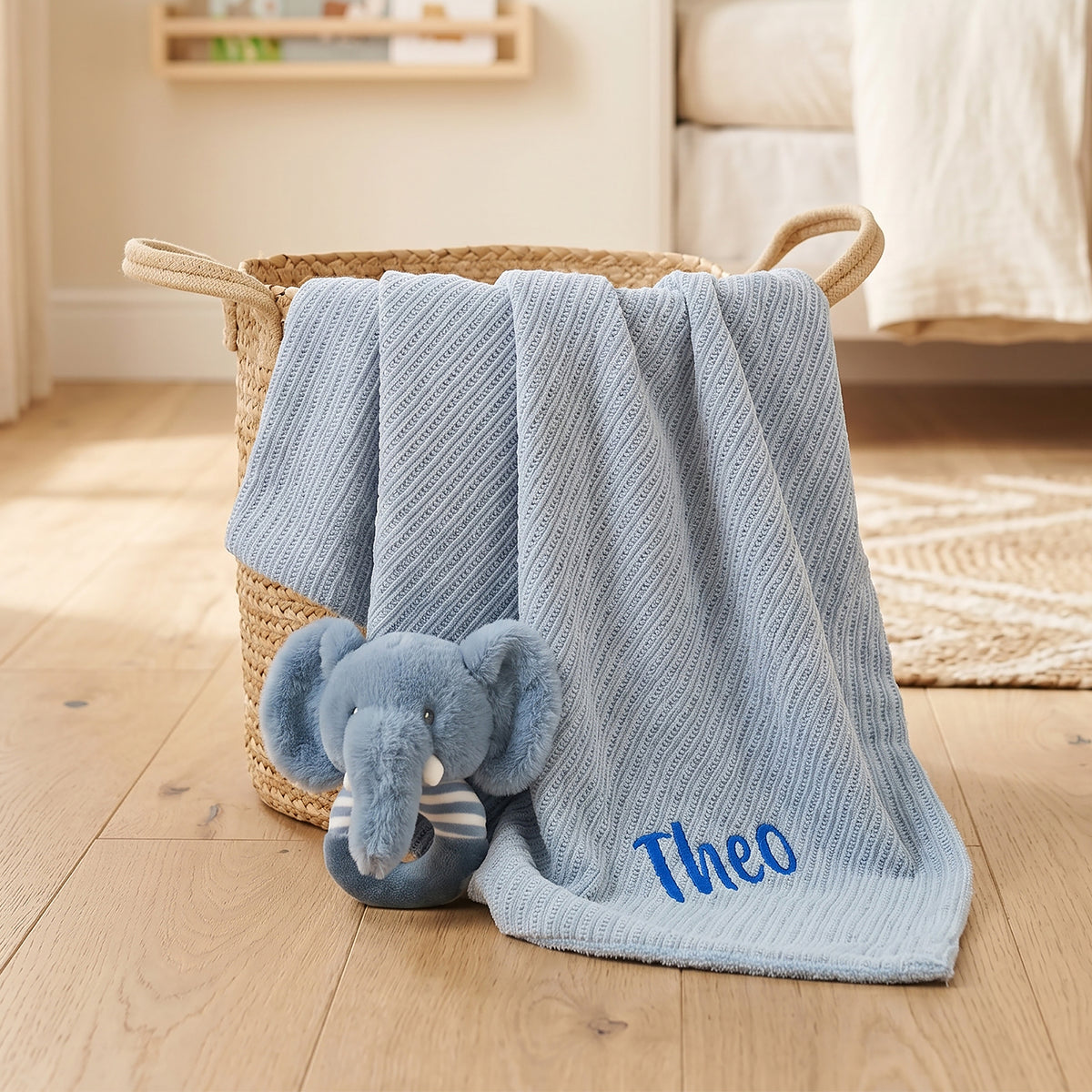A light blue textured baby blanket with the name 'Theo' embroidered in blue, draped over a woven basket next to a plush blue elephant toy in a softly lit nursery setting.