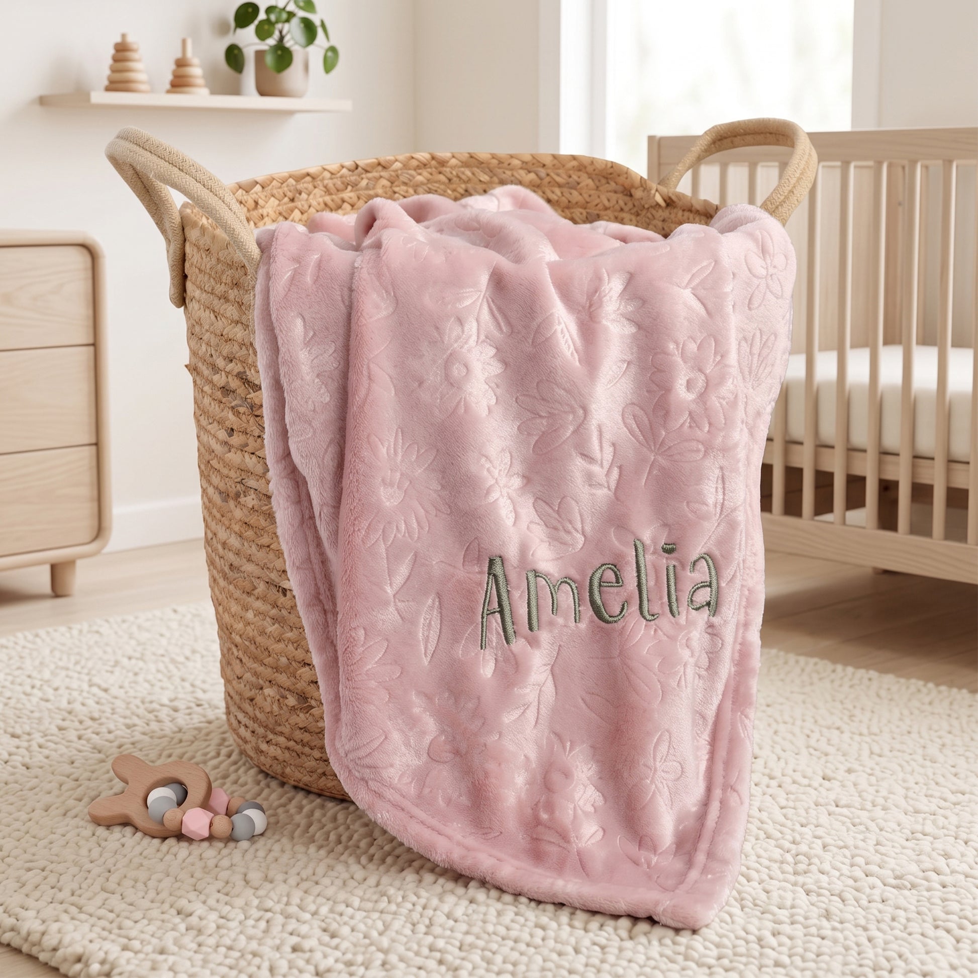 A soft pink plush blanket with an embossed floral pattern and the name 'Amelia' embroidered in green, draped over a woven wicker basket in a bright nursery setting.