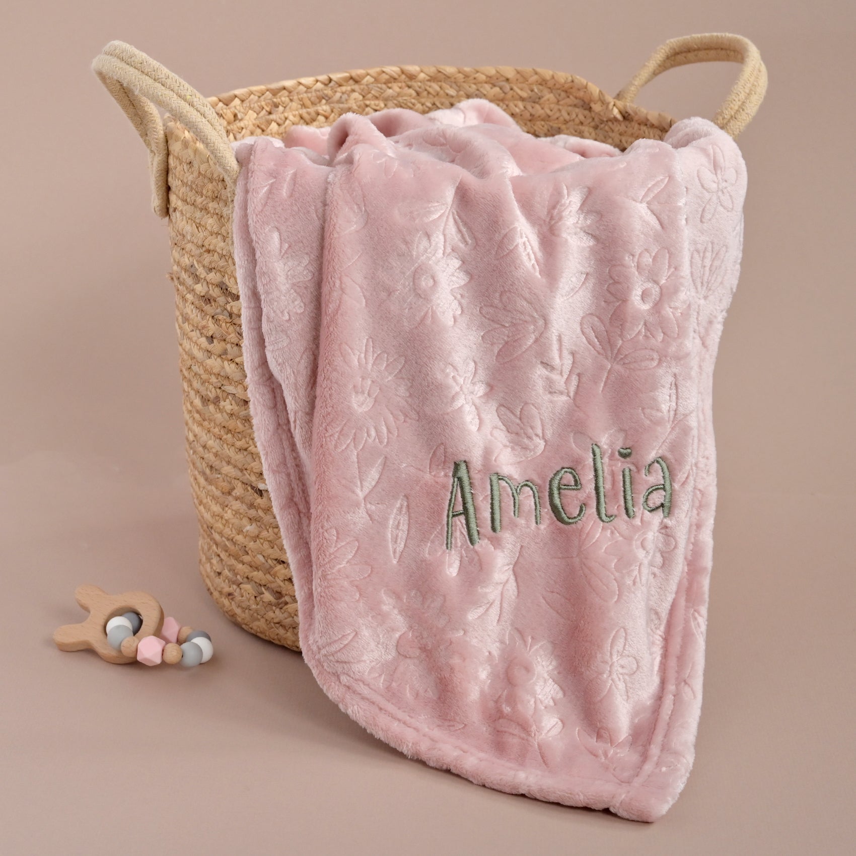 A soft pink plush blanket with an embossed floral pattern and the name 'Amelia' embroidered in green, draped over a woven straw basket on a beige background.