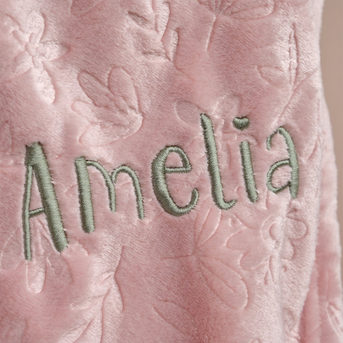 Close-up of a pink textured fabric with floral quilting and the name 'Amelia' embroidered in light green thread.