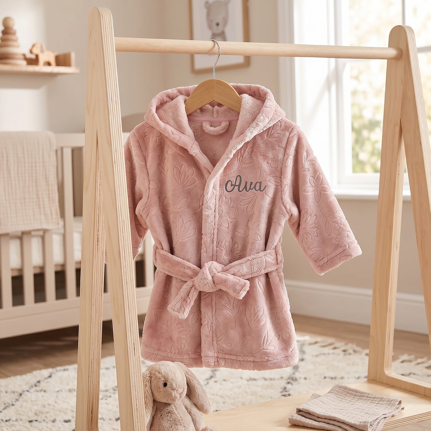 A pink plush hooded baby robe with a tie belt hangs on a wooden clothing rack in a softly lit nursery. The robe has an allover tonal floral texture and the name 'Ava' embroidered in dark script on the chest.