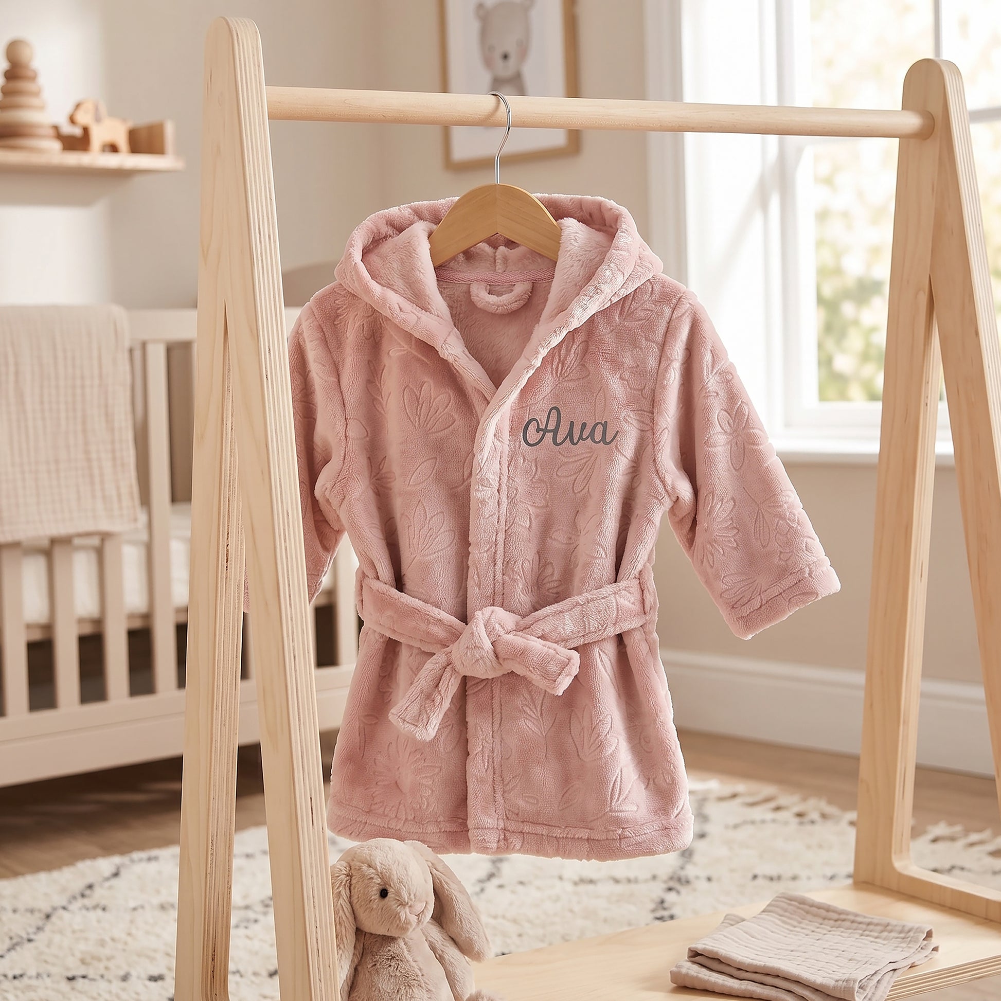 A pink plush hooded baby robe with a tie belt hangs on a wooden clothing rack in a softly lit nursery. The robe has an allover tonal floral texture and the name 'Ava' embroidered in dark script on the chest.