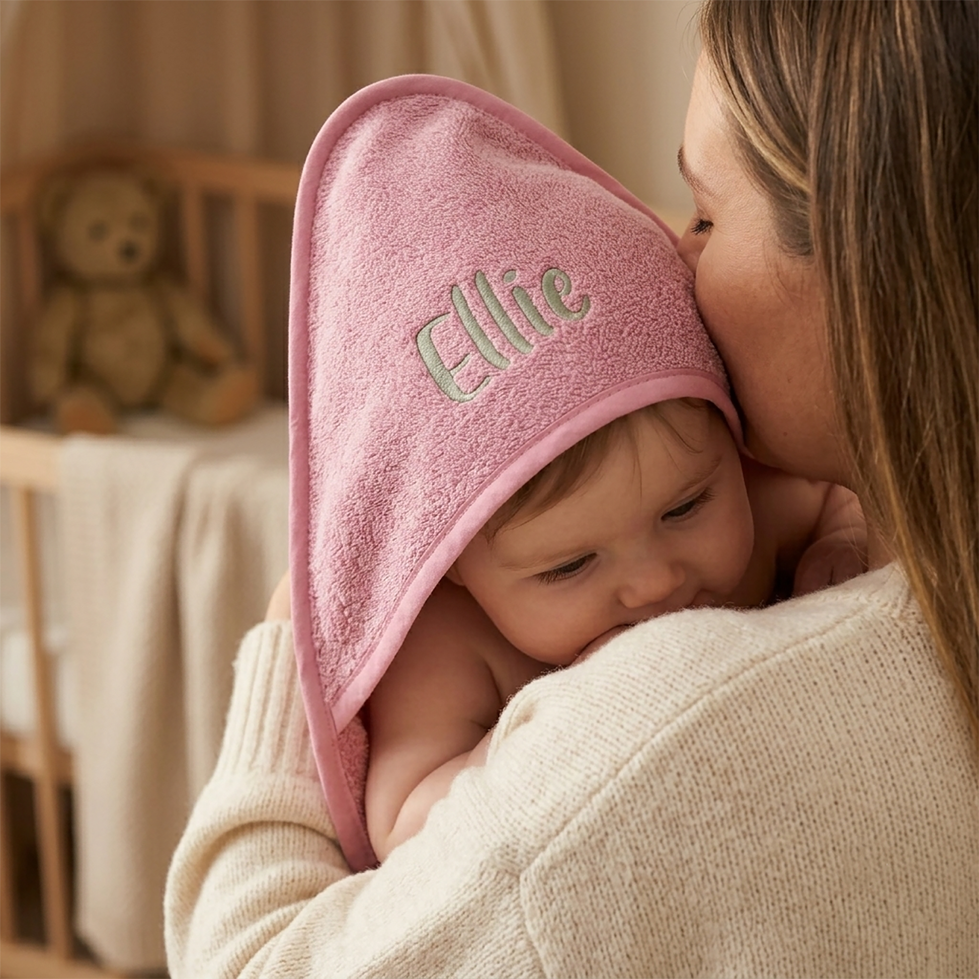 Dusty pink hooded baby towel in cotton terry, personalised with the name Ellie embroidered in sage green, tucked against a mother's shoulder.