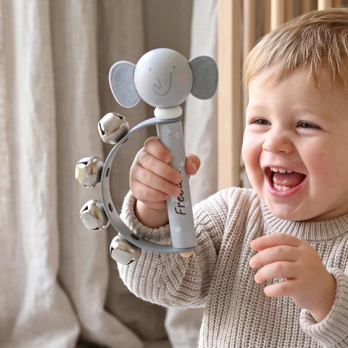 A smiling toddler in a beige knit sweater holds a gray elephant-shaped baby rattle toy with silver bells in front of a soft beige curtain background.