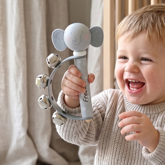 A smiling toddler in a beige knit sweater holds a gray elephant-shaped baby rattle toy with silver bells in front of a soft beige curtain background.