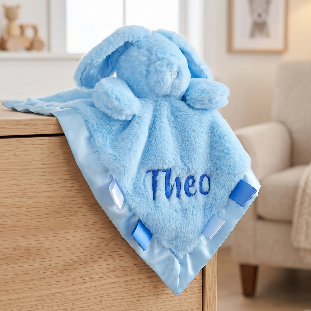 A light blue plush bunny lovey blanket with the name 'Theo' embroidered on it, draped over a wooden dresser in a softly lit room.