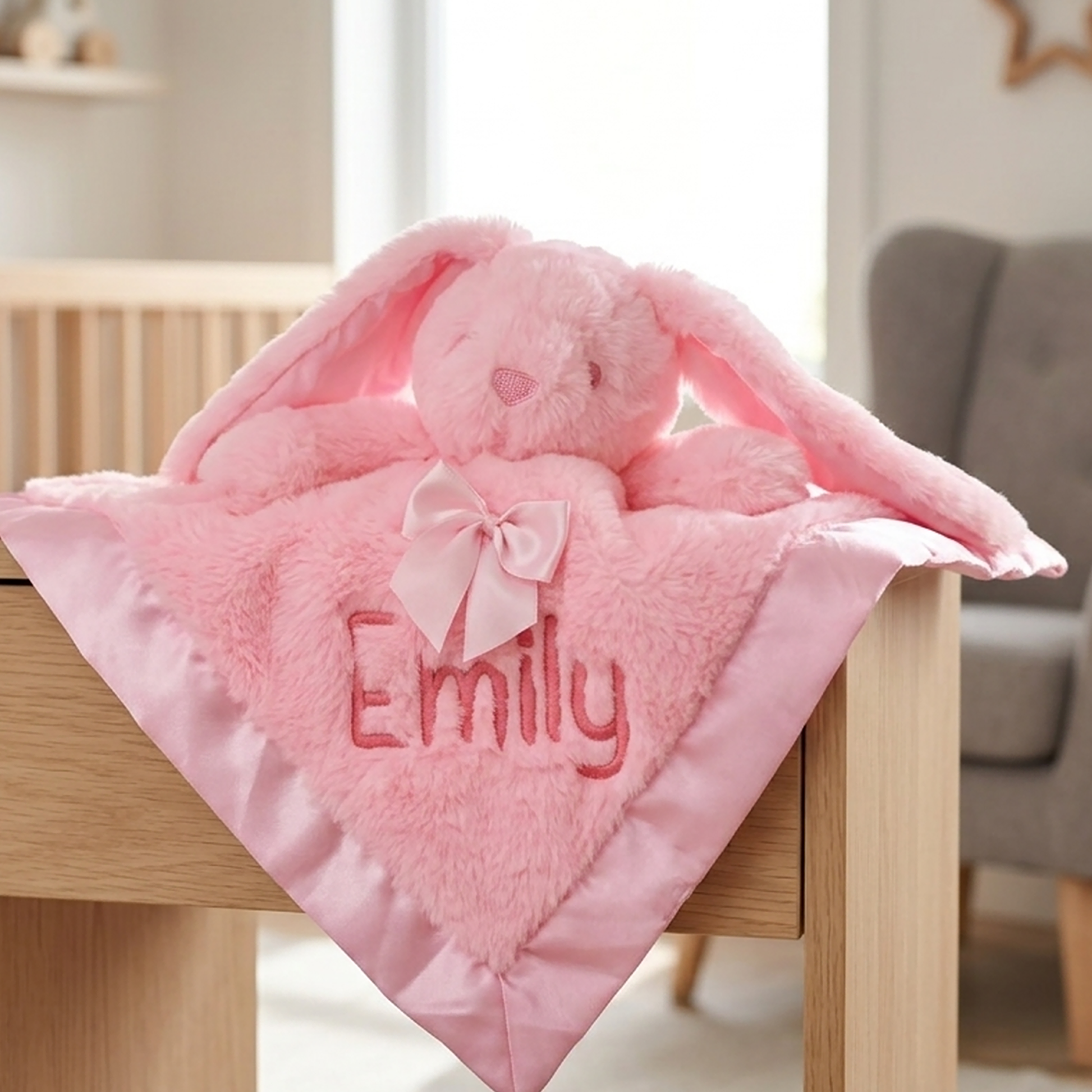 A pink plush bunny lovey blanket with long floppy ears, a satin border, and the name 'Emily' embroidered on the front, draped over a wooden table in a softly lit nursery setting.