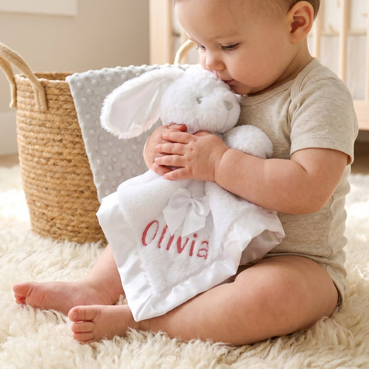 Baby cuddling a white faux fur bunny comforter with satin trim and bow, personalised with the name "Olivia" in red embroidery, next to a woven basket.