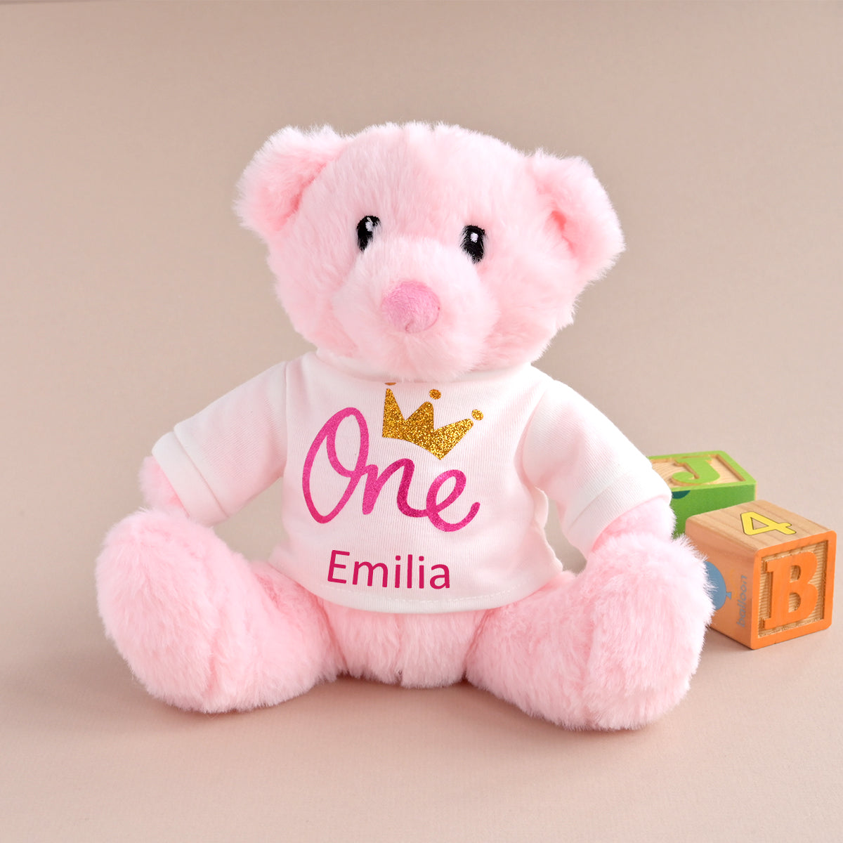 A pink plush teddy bear wearing a white shirt with 'One' in pink script, a gold glitter crown, and the name 'Emilia' in red, sitting next to wooden toy blocks on a beige background.
