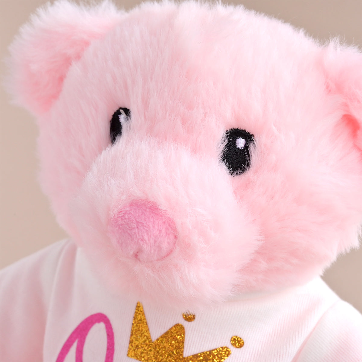 A close-up of a pink plush pig toy wearing a white shirt with a gold glitter crown graphic and pink lettering, set against a soft beige background.