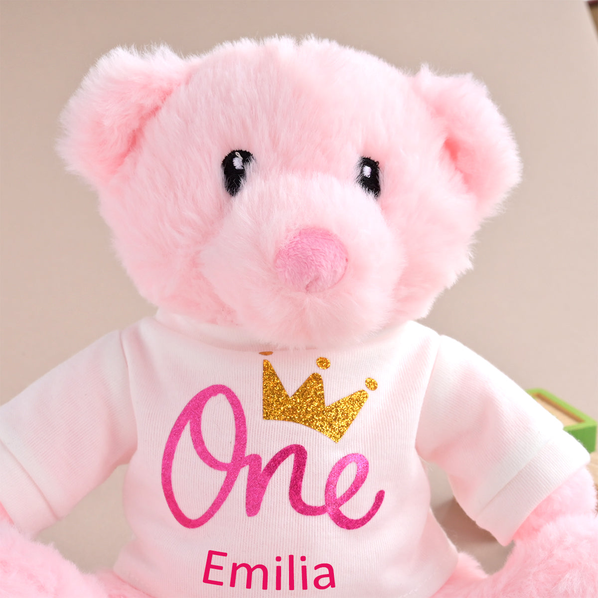 A pink plush teddy bear wearing a white long-sleeve shirt with a glittery gold crown and the words 'One' and 'Emilia' in pink, photographed against a soft beige background.