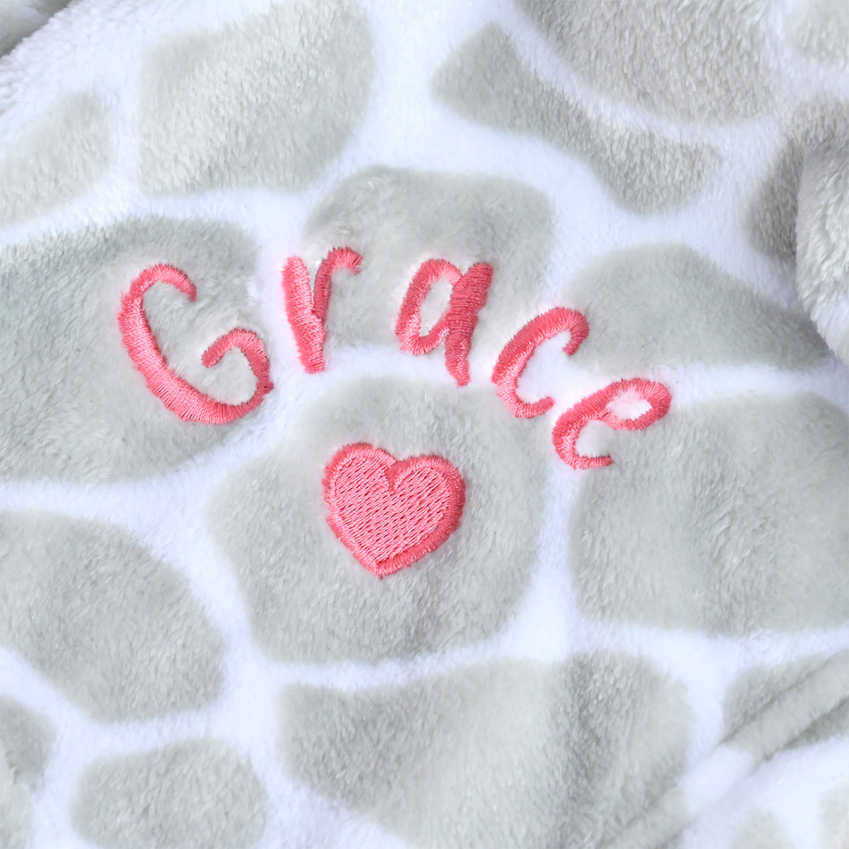 Close-up of a plush fleece blanket with a light gray and white giraffe print pattern, featuring the name 'Grace' and a pink heart embroidered in the center.