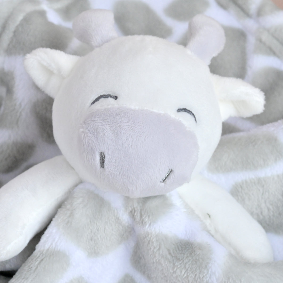 A close-up of a plush giraffe comforter or stuffed toy with a soft white face and neck, gray spots, and embroidered closed eyes on a matching gray-and-white spotted blanket.