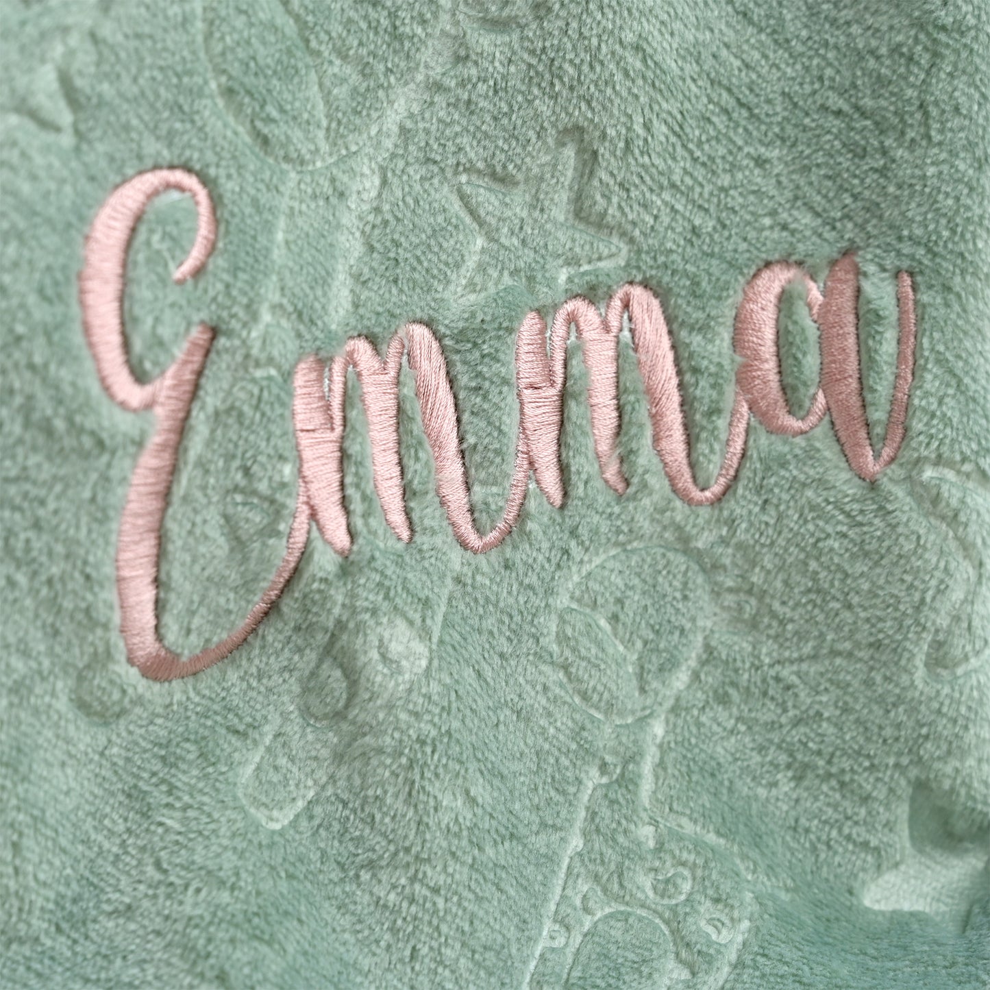 Close-up of a personalized plush blanket featuring the name 'Emma' embroidered in light pink cursive thread over a textured mint green fabric background with subtle character designs.