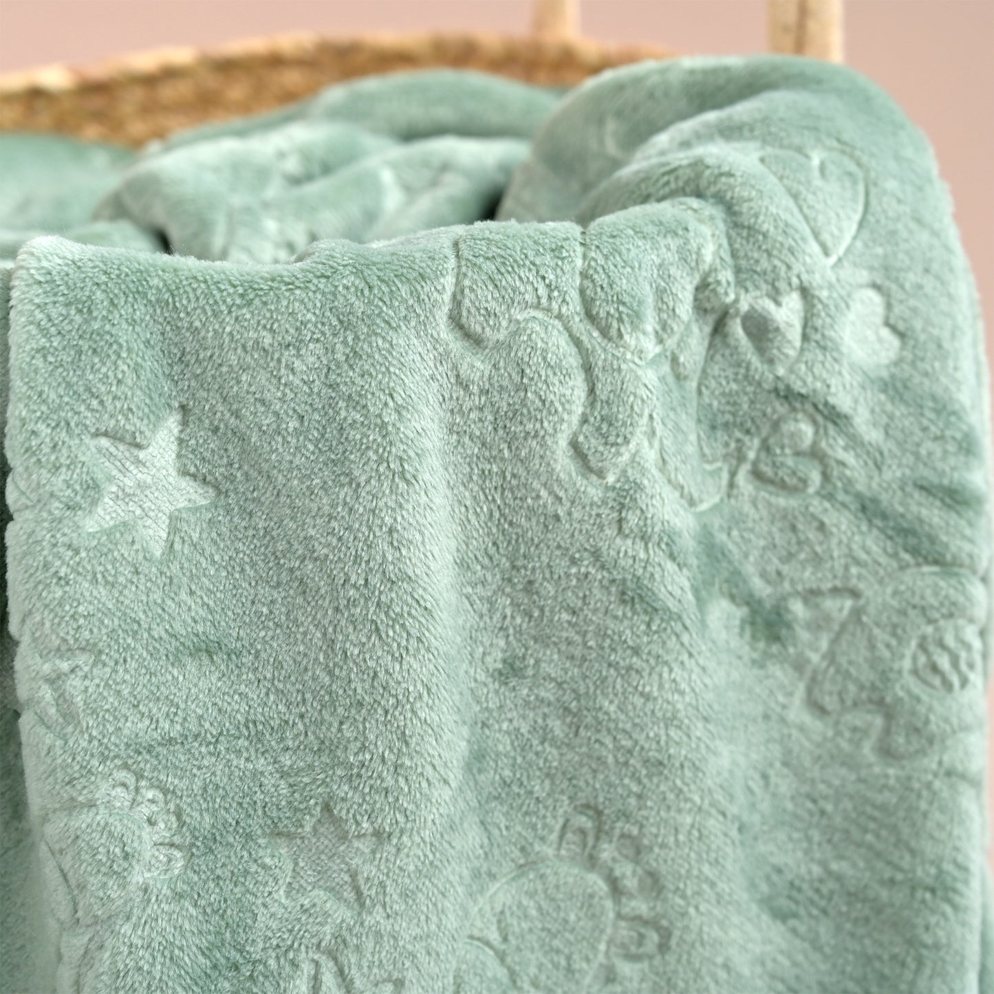 A close-up of a soft, plush baby blanket in a dusty mint green color, featuring embossed animal and star patterns, draped over a woven basket.