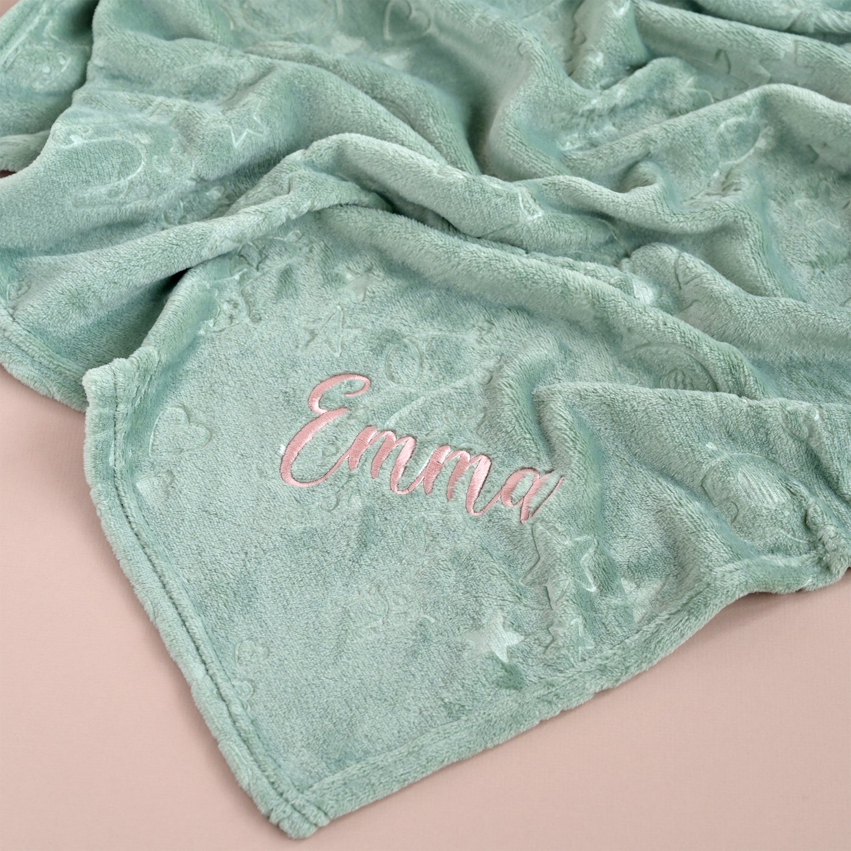 A mint green plush blanket with a textured star pattern, featuring the name 'Emma' embroidered in pink script on one corner, laid out on a soft pink background.