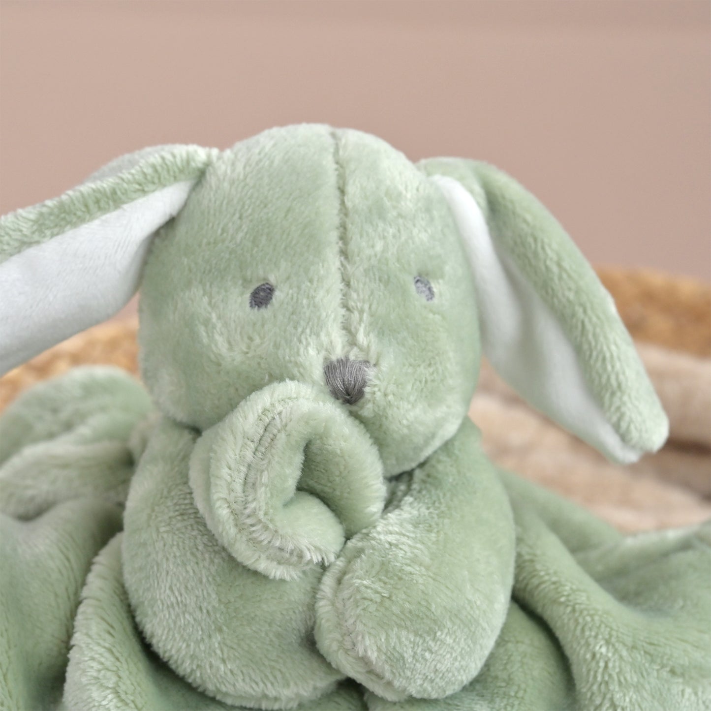 A close-up of a soft plush bunny blanket in light sage green with white inner ears and a gray embroidered nose and eyes, resting on a neutral surface.