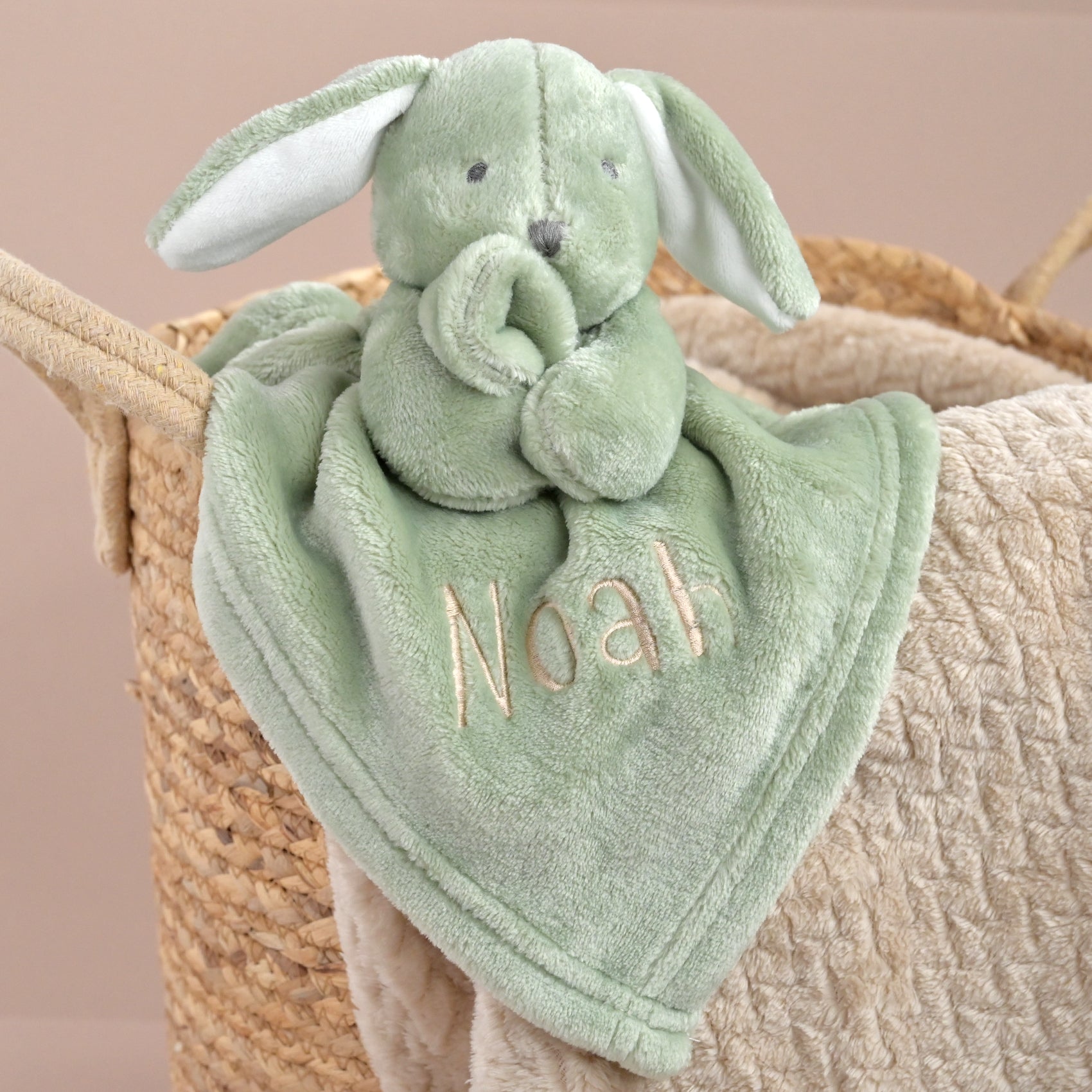 A personalized plush bunny lovey blanket in sage green, featuring a soft plush bunny head with floppy ears and a matching square blanket embroidered with the name 'Noah' in a light beige font. The toy is styled inside a woven straw baby basket with a beige quilted blanket.