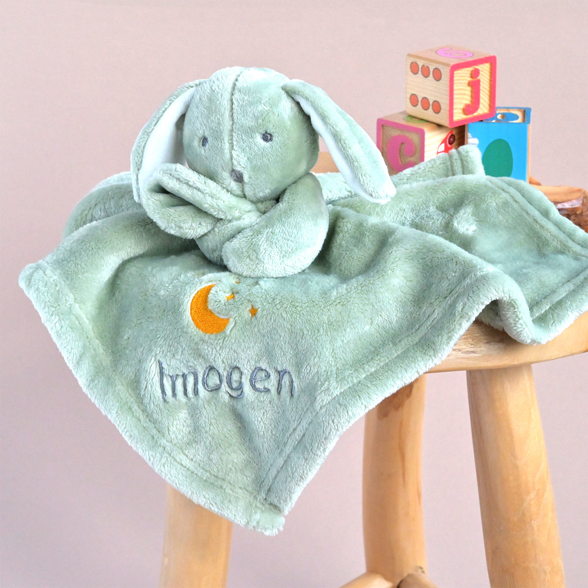 A personalized baby security blanket featuring a plush bunny head and a soft green blanket with an embroidered crescent moon and the name 'Imogen'. The blanket is draped over a wooden stool next to colorful toy blocks against a solid pink background.