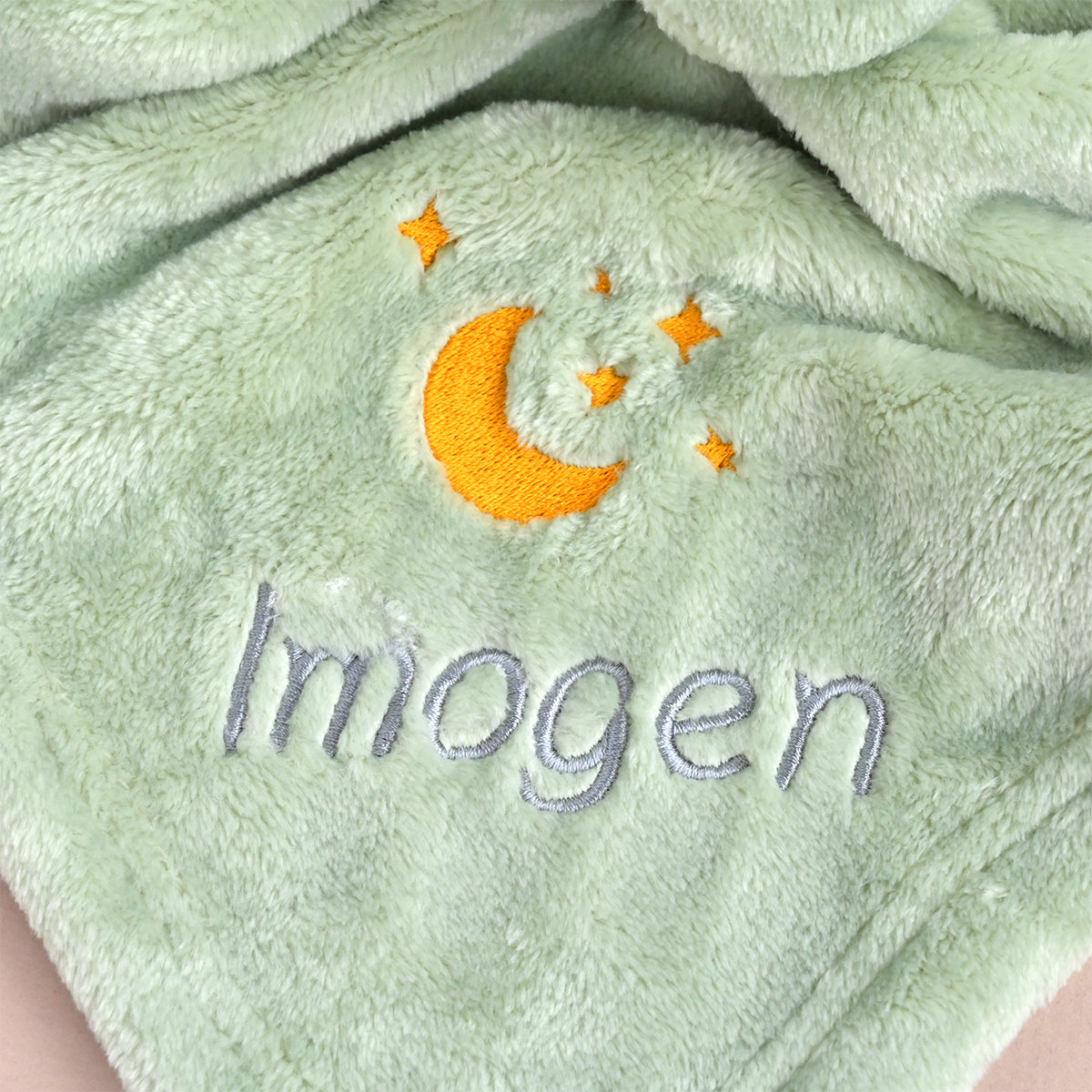 A close-up of a plush mint green blanket featuring an embroidered orange crescent moon and stars, with the name 'Imogen' stitched in gray below.