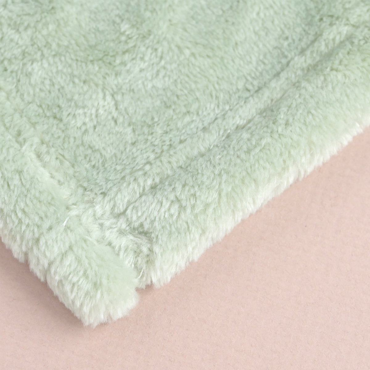 A close-up of a soft, pale mint green plush or sherpa fabric corner with a folded over edge, set against a solid light pink background.