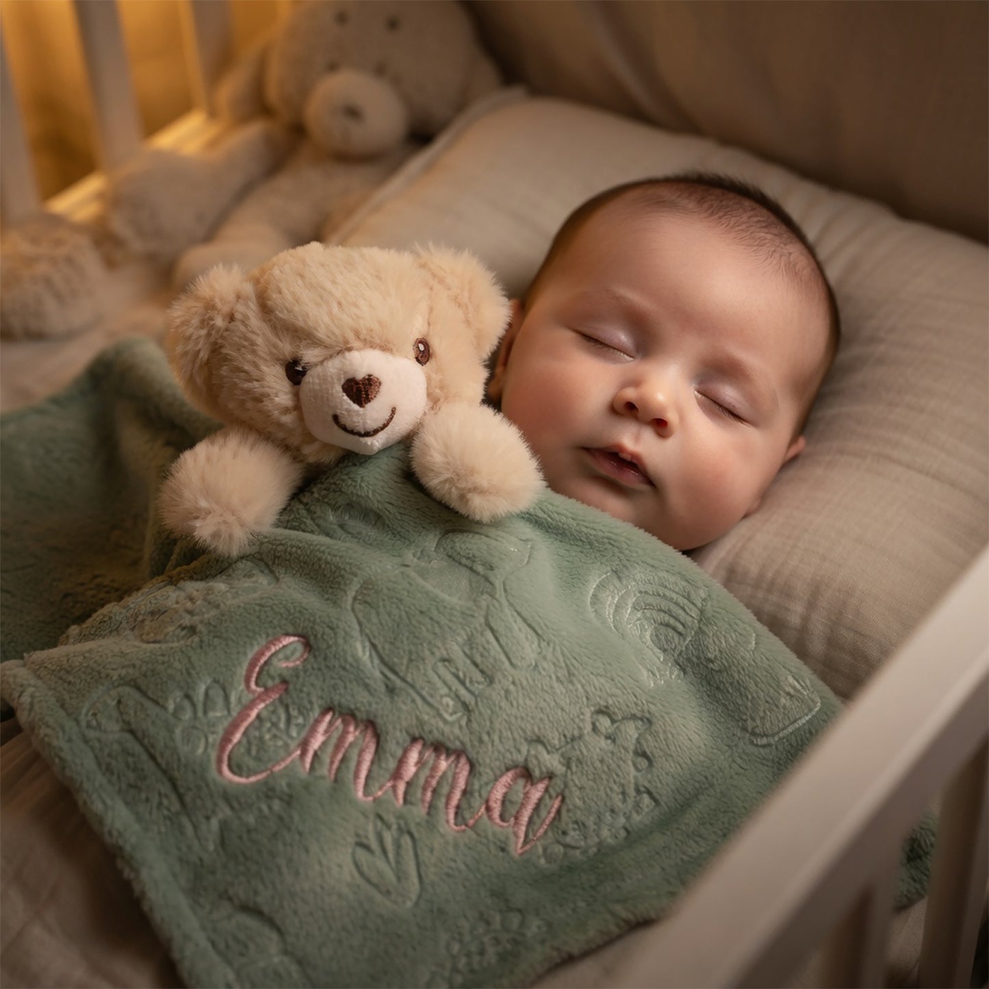 A sleeping baby tucked in a crib, wrapped in a personalized sage green blanket with the name 'Emma' embroidered in pink, holding a small beige teddy bear.