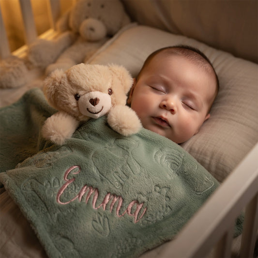 A sleeping baby tucked in a crib, wrapped in a personalized sage green blanket with the name 'Emma' embroidered in pink, holding a small beige teddy bear.