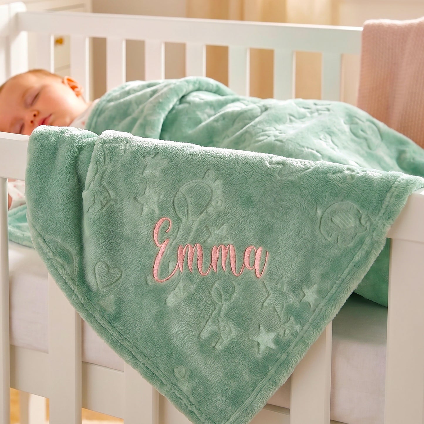 A sleeping baby tucked in a white crib, wrapped in a mint green plush blanket with embossed stars and the name 'Emma' embroidered in pink script.