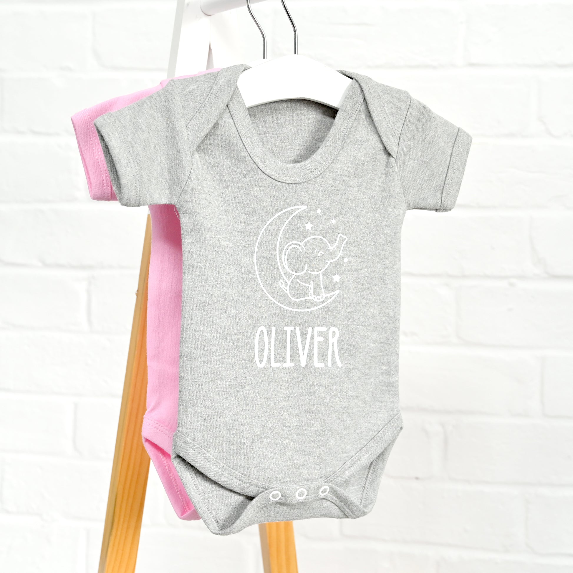 A heather gray short-sleeve baby bodysuit hanging on a white hanger, featuring a white line-art graphic of a baby elephant on a crescent moon with the name 'OLIVER' printed below. A pink bodysuit is partially visible behind it on a wooden hanger stand against a white brick wall background.