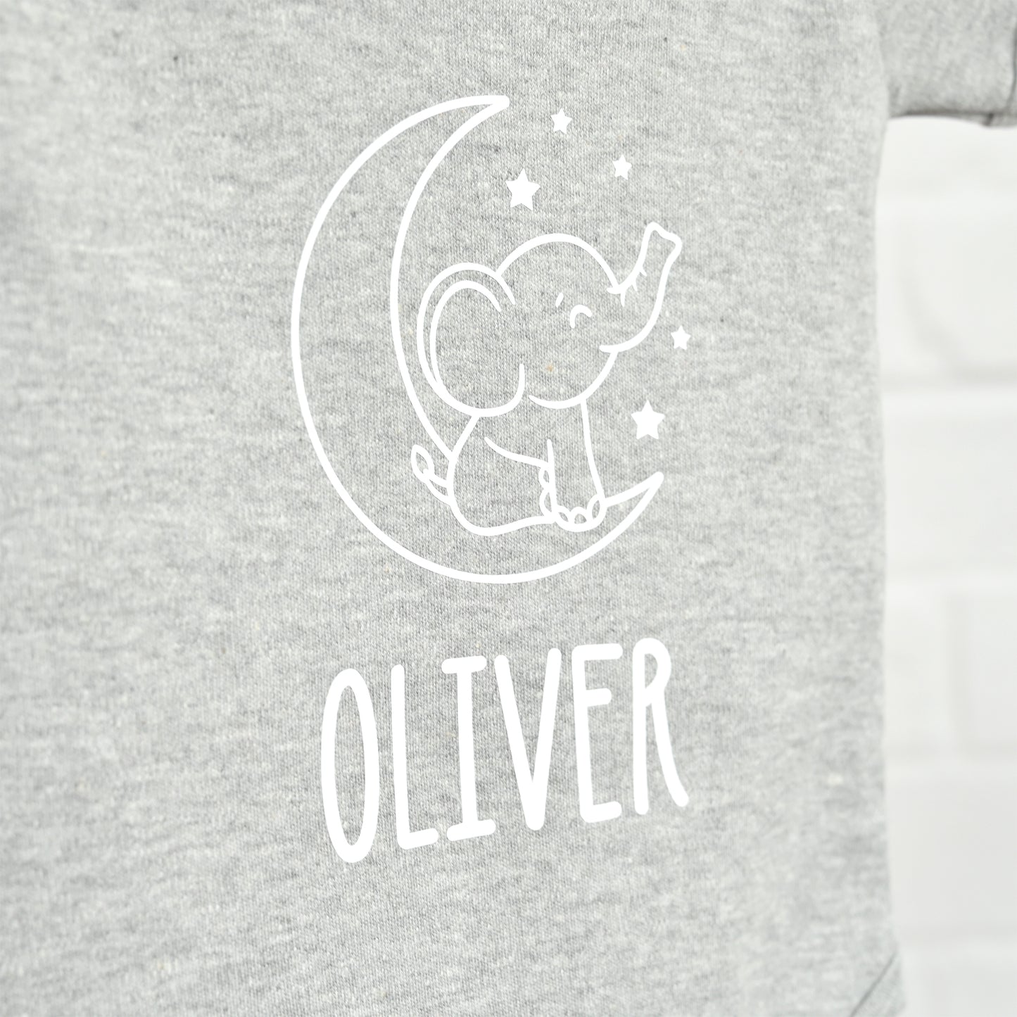 Close-up of a heather gray garment with a white line-art graphic of an elephant sitting on a crescent moon and stars, with the name 'OLIVER' printed below in white.
