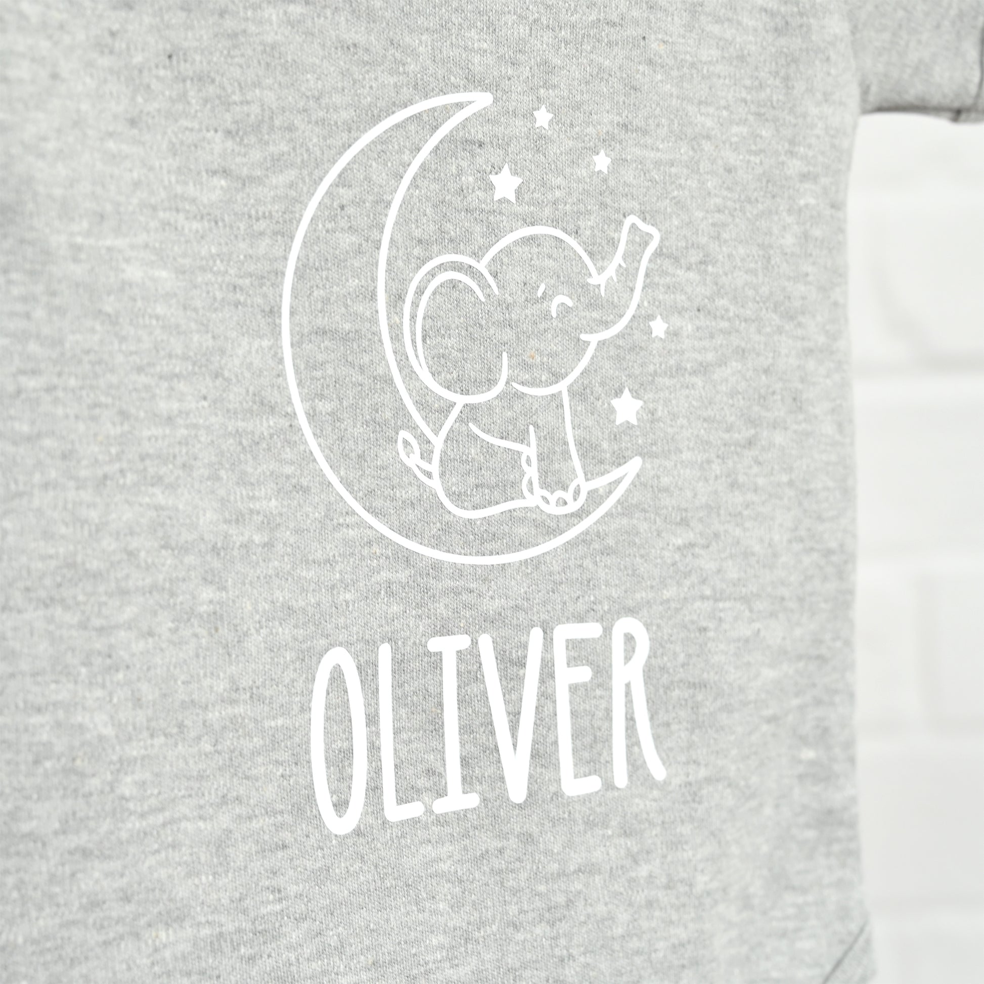 Close-up of a heather gray garment with a white line-art graphic of an elephant sitting on a crescent moon and stars, with the name 'OLIVER' printed below in white.