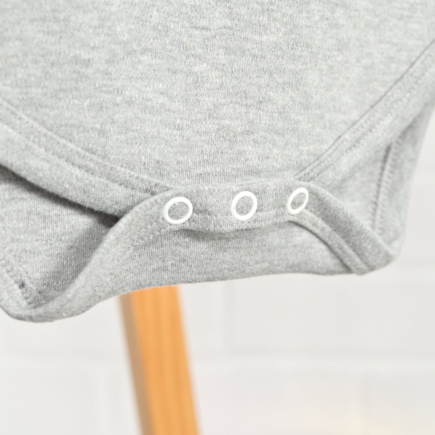 Close-up of a light gray heather baby bodysuit or romper with three white snap closures at the crotch, hanging on a wooden stand against a light background.